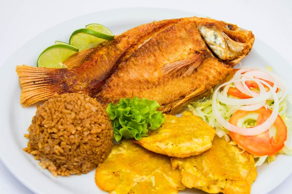 Delicious fried fish served with seasoned rice, tostones, and fresh salad on a white plate.
